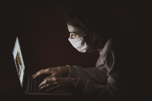 Eine Frau mit Mundschutz, die auf einen Laptop schaut und etwas tippt. Der Hintergrund ist düster.