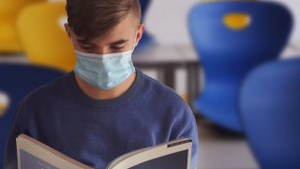 Jugendlicher mit Mund-Nase-Schutz ließt ein Buch. Im Hintergrund stehen leere Stühle.