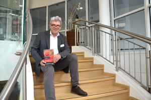 Prof. Bering sitzt auf einer Treppe und hält das Buch in den Händen.